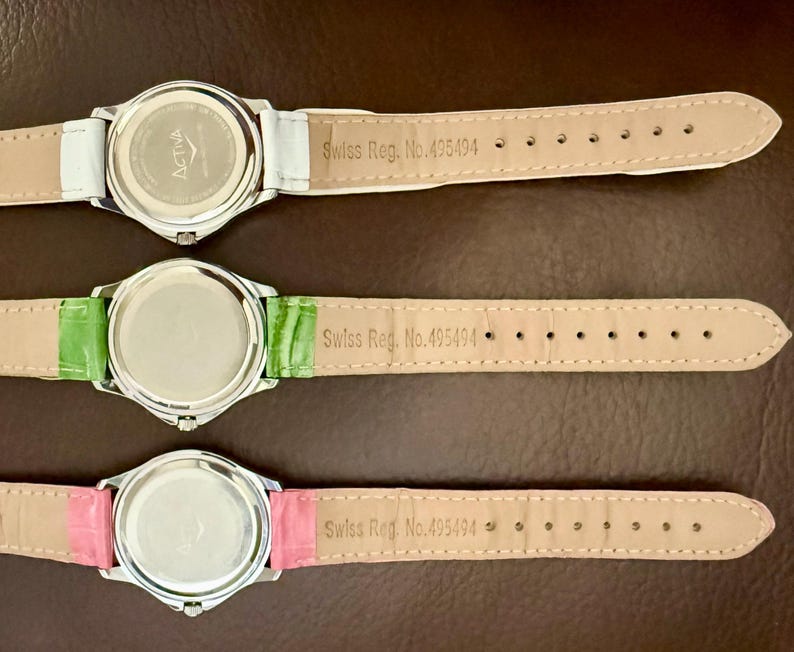 Op de afbeelding: Drie horloges met zilverkleurige kasten en leren bandjes in wit, groen en roze. De achterkanten van de horloges zijn zichtbaar, met de merknaam "ACTIVA" en de tekst "Swiss Reg. No. 495494" op de bandjes.