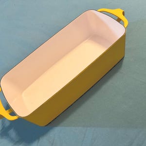 May include: A rectangular baking dish with a bright yellow exterior and a smooth white interior. The vessel features two yellow handles on opposite ends and a black rim along the top edge, providing a clean and functional design for kitchen use.