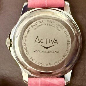 Op de afbeelding: Een roestvrijstalen horloge met een roze leren band. De achterkant van het horloge toont de merknaam "Activa", modelnummer SL233-002 en tekst die Zwitserse onderdelen en waterbestendigheid aangeeft.