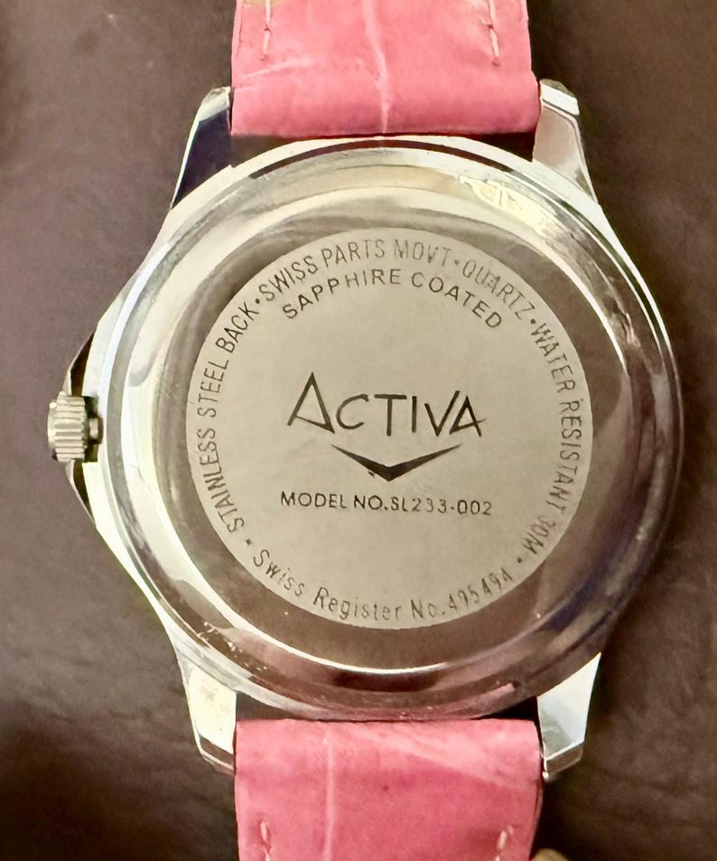 Op de afbeelding: Een roestvrijstalen horloge met een roze leren band. De achterkant van het horloge toont de merknaam "Activa", modelnummer SL233-002 en tekst die Zwitserse onderdelen en waterbestendigheid aangeeft.