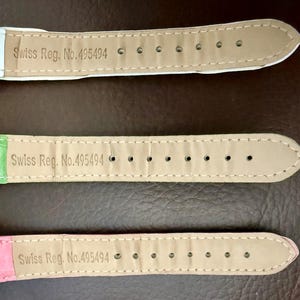 Op de afbeelding: Drie horlogebandjes in pastelkleuren: wit, groen en roze. Elk bandje heeft de tekst "Swiss Reg. No. 495494" erop gedrukt. De bandjes zijn gemaakt van een textuurrijk materiaal en hebben meerdere gaatjes.