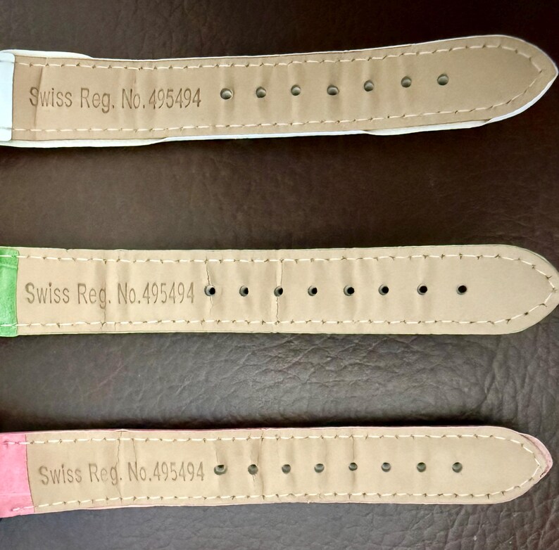 Op de afbeelding: Drie horlogebandjes in pastelkleuren: wit, groen en roze. Elk bandje heeft de tekst "Swiss Reg. No. 495494" erop gedrukt. De bandjes zijn gemaakt van een textuurrijk materiaal en hebben meerdere gaatjes.