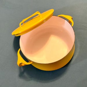 May include: A yellow enameled cast iron Dutch oven with a matching lid. The interior is light pink. The lid has a handle, and the pot has two side handles. The pot is sitting on a blue-green surface.