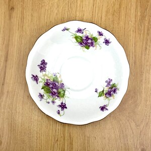 May include: White saucer with a scalloped edge and a gold rim. The saucer is decorated with clusters of purple violets and green leaves. A delicate floral design for serving tea or coffee.