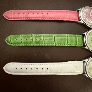 Op de afbeelding: Drie polshorloges met leren bandjes in roze, groen en wit. De wijzerplaten van de horloges zijn zilverkleurig met bijpassende gekleurde wijzerplaten. De bandjes hebben een textuur, krokodilachtig patroon. De horloges worden gepresenteerd op een bruin oppervlak.
