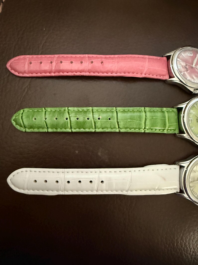 Op de afbeelding: Drie polshorloges met leren bandjes in roze, groen en wit. De wijzerplaten van de horloges zijn zilverkleurig met bijpassende gekleurde wijzerplaten. De bandjes hebben een textuur, krokodilachtig patroon. De horloges worden gepresenteerd op een bruin oppervlak.