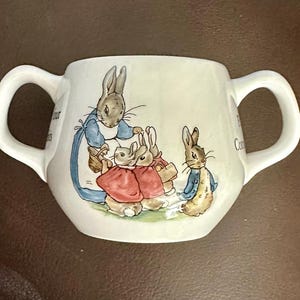 May include: White ceramic mug with two handles, featuring a colorful illustration of rabbits in a storybook scene. The rabbits are dressed in clothing, and the mug has text on the side.
