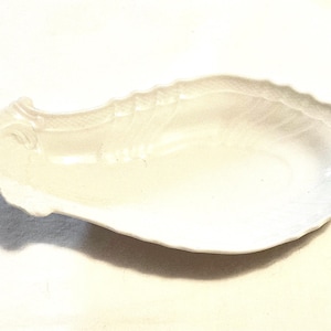 May include: A white, oval-shaped ceramic serving dish with a scalloped edge. The dish has a smooth, glossy surface and a subtle, textured design. The dish is ideal for serving appetizers or small dishes.