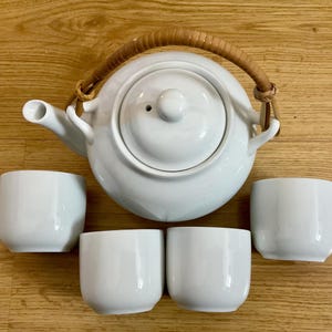May include: A white ceramic teapot with a woven handle and four matching teacups. The teapot has a rounded shape and a spout. The teacups are small and cylindrical. The set is arranged on a wooden surface.