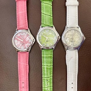 Op de afbeelding: Drie horloges met bijpassende bandjes in roze, groen en wit. Elk horloge heeft een zilverkleurige kast en een leren bandje. De wijzerplaten tonen de cijfers 3, 6, 9 en 12. De bandjes hebben een textuur met krokodillenpatroon.