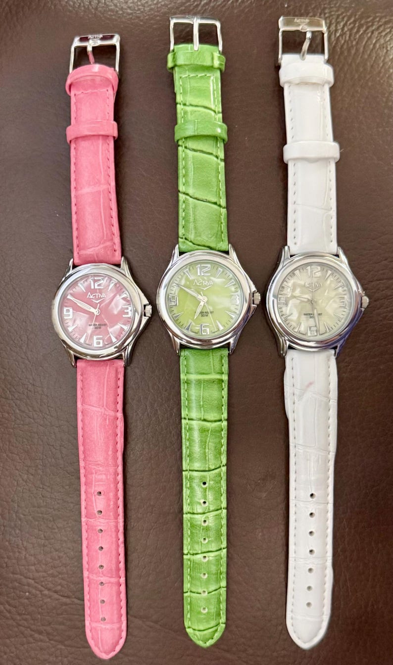 Op de afbeelding: Drie horloges met bijpassende bandjes in roze, groen en wit. Elk horloge heeft een zilverkleurige kast en een leren bandje. De wijzerplaten tonen de cijfers 3, 6, 9 en 12. De bandjes hebben een textuur met krokodillenpatroon.