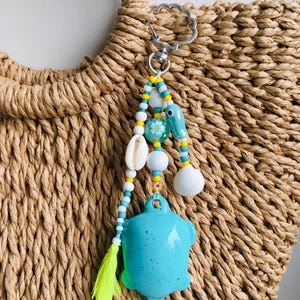 Puede incluir: Un llavero con un dije de tortuga turquesa con una borla amarilla, conchas marinas y cuentas de colores. El llavero está sujeto a un cierre plateado, colgando de un bolso de paja tejido. Las cuentas son blancas, amarillas y azules.