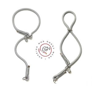 Puede incluir: Dos correas de cuerda color gris plateado para perros, con construcción de cordón trenzado, terminales metálicos y topes ajustables de aspecto de cuero. Entre las correas hay un logotipo circular con el texto BARKING MAD DOGGY WEAR.
