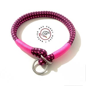May include: A round dog collar made of braided dark purple and bright pink rope, featuring two sections wrapped in vibrant pink cord and two silver metal rings. The center of the collar displays a logo with the text BARKING MAD DOGGY WEAR.