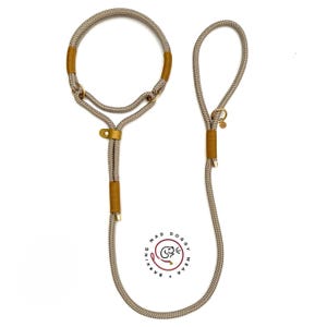 May include: A beige rope slip lead for pets featuring mustard yellow thread accents, gold-toned metal rings, and a circular logo with the text BARKING MAD DOGGY WEAR. The leash includes a fixed loop handle and an adjustable collar section.