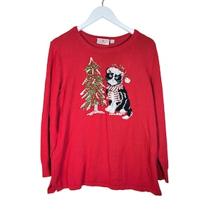May include: A red long-sleeve sweater featuring a sequined Christmas tree and a black and white cat wearing a Santa hat and scarf. The sweater is hanging on a white hanger.