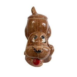 May include: A brown ceramic dog-shaped cookie jar with a lid. The dog has a long snout, floppy ears, and a red tongue sticking out. The eyes have white paint that is chipped. The jar is a vintage item.