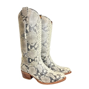 May include: A pair of tall, knee-high cowboy boots with a snakeskin pattern in shades of gray, black, and cream. The boots have a pointed toe, a stacked heel, and a leather sole. The boots are made of a snakeskin material.