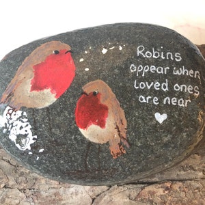 May include: A painted rock with two robins on it. The rock has the text "Robins appear when loved ones are near" written on it in white paint.