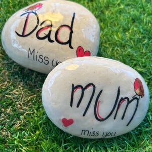 May include: Two painted rocks with the words "Dad" and "Mum" written on them. Each rock has a small red heart and a robin painted on it. The words "Miss you" are written on the "Dad" rock and "Miss you" is written on the "Mum" rock.