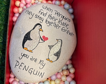 May include: A white painted rock with two penguins and a red heart. The rock is in a red box with pink and blue packing peanuts. The text on the rock reads "When penguins find their mate, they stay together forever. You are my penguin."