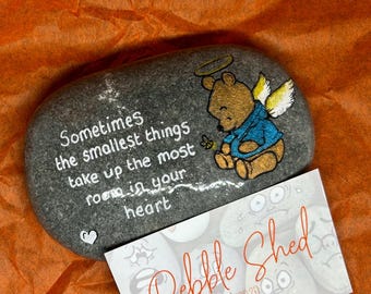 May include: Hand-painted grey stone featuring a Winnie the Pooh character with angel wings and a halo. The stone has the text "Sometimes the smallest things take up the most room in your heart". The stone is resting on a white card with the text "Pebble Shed est 2020" and social media details.