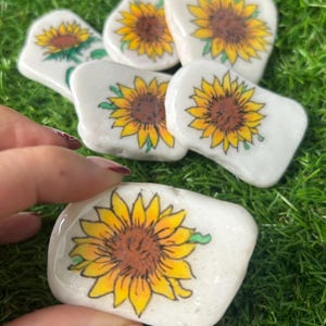 Puede incluir: Un conjunto de piedras blancas y lisas, cada una pintada a mano con un diseño de girasol vibrante. Los girasoles presentan pétalos amarillos y centros marrones, con detalles verdes. Las piedras varían en tamaño y forma, ideales para decoración.