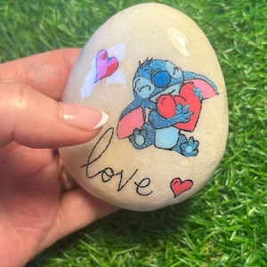 May include: A painted rock featuring a blue cartoon character holding a red heart. The rock also has the word "love" written on it and several pink hearts.