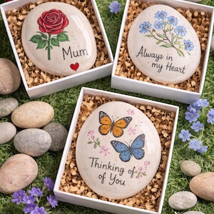 May include: Three white gift boxes containing smooth, oval-shaped stones. One stone features a red rose and the text 'Mum' with a heart. Another shows blue flowers and 'Always in my Heart'. The third displays two butterflies and 'Thinking of You'.