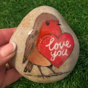 May include: A painted stone with a robin design holding a red heart with the words "love you". The bird has a red breast and brown wings. The stone is beige and has a smooth texture. The artwork is on a natural stone.