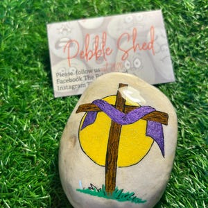 May include: A painted rock with a brown cross and a purple ribbon wrapped around it. The cross is in front of a yellow sun. The rock is on a green grassy surface. The text "Pebble Shed" is visible in the background.