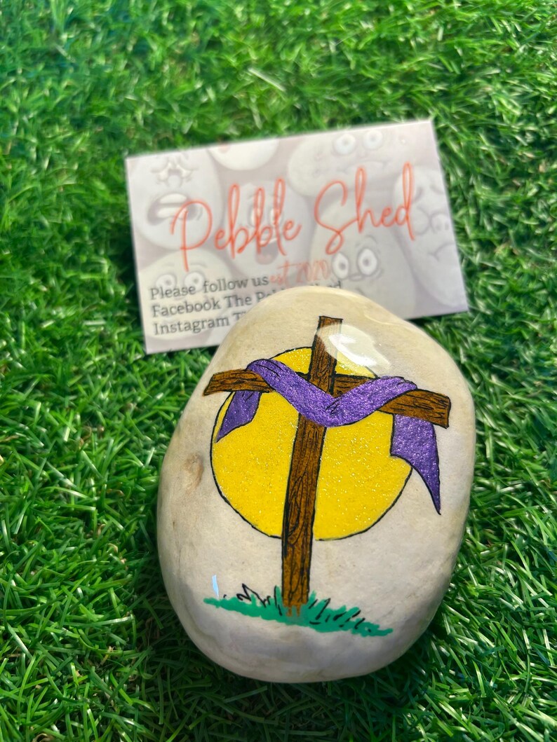 May include: A painted rock with a brown cross and a purple ribbon wrapped around it. The cross is in front of a yellow sun. The rock is on a green grassy surface. The text "Pebble Shed" is visible in the background.