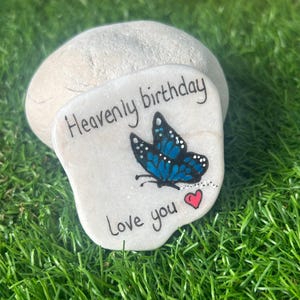 Può includere: Una pietra dipinta con le parole "Heavenly birthday Love you" e un disegno di farfalla blu. Un piccolo cuore rosso è anche dipinto sulla pietra. La pietra è appoggiata su un letto di erba verde.