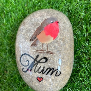 May include: A painted stone featuring a robin with a red breast and the word "Mum" in cursive, with a small red heart. The stone is set against a backdrop of green grass.