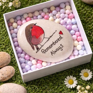 Op de afbeelding: Een beschilderde steen met een afbeelding van een roodborstje en de tekst "Remembered Always". De steen ligt in een witte doos gevuld met pastelkleurige bollen. De scène wordt gecompleteerd door madeliefjes en kiezels op groen gras.