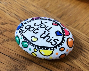 May include: A white painted rock with a black circle around the words "You got this" in black lettering. The rock is decorated with colorful dots and hearts.