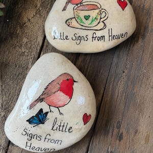 May include: Two smooth, cream-colored stones painted with illustrations. One stone features a robin perched on a teacup with a red heart and the text "Little Signs from Heaven." The other stone shows a robin and a blue butterfly with a red heart and the text "little Signs from Heaven."