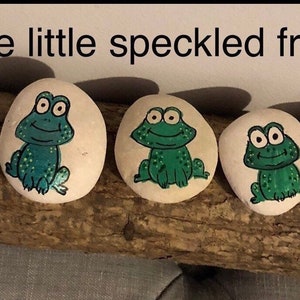 Könnte beinhalten: Fünf bemalte Steine mit Froschillustrationen. Jeder Stein zeigt ein anderes Froschdesign in Grün und Blau, mit dem Text "Five little speckled frogs" darüber. Die Steine sind auf einem Holzstück angeordnet.