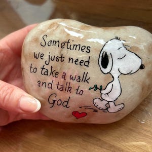 May include: A heart-shaped, painted stone with the text "Sometimes we just need to take a walk and talk to God." A cartoon Snoopy character is walking with a sprig of leaves. A red heart is painted below the text.