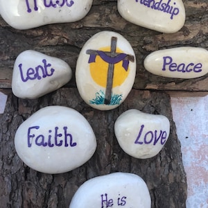 May include: Painted white rocks with purple lettering spelling out the words "Trust", "Friendship", "Lent", "Peace", "Faith", "Love", and "He is risen!"  A painted rock with a yellow sun and a purple cross is in the center.