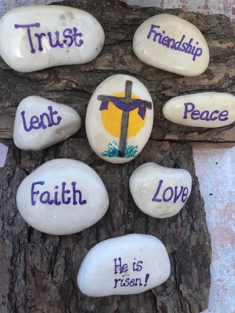 May include: Painted white rocks with purple lettering spelling out the words "Trust", "Friendship", "Lent", "Peace", "Faith", "Love", and "He is risen!"  A painted rock with a yellow sun and a purple cross is in the center.