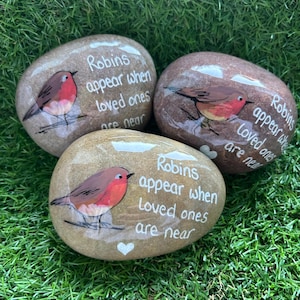 May include: Three painted stones featuring a robin design and the text "Robins appear when loved ones are near." The robins have red breasts and brown wings. The stones are displayed on a bed of green grass.