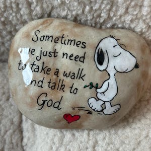 May include: A painted stone with the text "Sometimes we just need to take a walk and talk to God." A cartoon Snoopy character is walking with a sprig of leaves. A red heart is painted below the text.