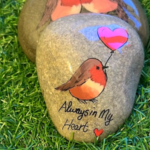 May include: Hand-painted decorative stones featuring robin bird designs. One stone has a robin holding a heart-shaped balloon, with the text "Always in My Heart." The other stones show two robins. The stones are on a bed of green grass.