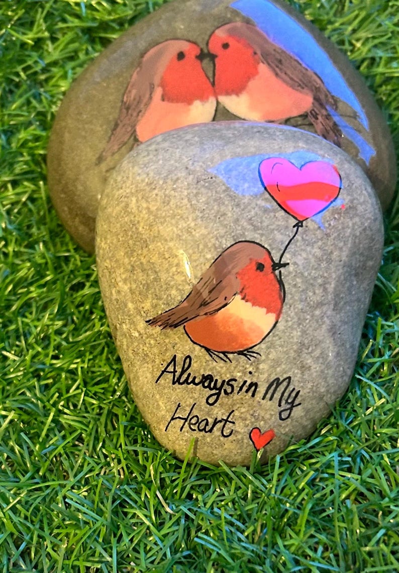 May include: Hand-painted decorative stones featuring robin bird designs. One stone has a robin holding a heart-shaped balloon, with the text "Always in My Heart." The other stones show two robins. The stones are on a bed of green grass.