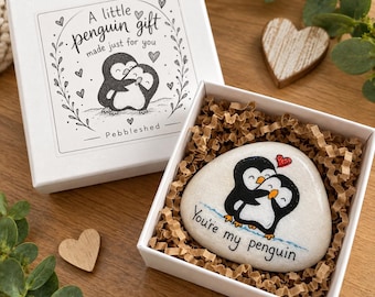 Puede incluir: Una caja de regalo blanca que contiene una piedra lisa y clara pintada con dos pingüinos blancos y negros abrazados bajo un pequeño corazón rojo. El texto en la piedra dice "You're my penguin". La tapa de la caja muestra una ilustración igual.