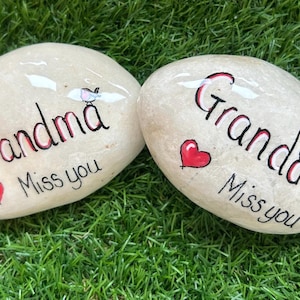 Może przedstawiać: Dwa gładkie, jasne kamienie rzeczne z ręcznie malowanym tekstem i ilustracjami. Na jednym kamieniu widnieje napis Grandma Miss you z czerwonym sercem i małym ptaszkiem. Na drugim kamieniu widnieje napis Grandad Miss you z czerwonym sercem i ptakiem.