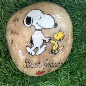 以下が含まれることがあります： スヌーピーとウッドストックが手をつないでいる手描きの滑らかな石。 下には「Best friends」の文字と赤いハートが描かれています。 背景は緑色です。