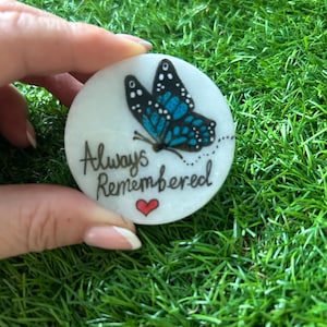 Może przedstawiać: Okrągły, biały kamień z niebiesko-czarnym motylem. Napis "Always Remembered" jest napisany czarnym pismem, z małym czerwonym sercem poniżej. Kamień jest trzymany na tle zielonej trawy.