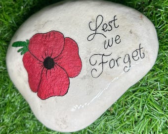 May include: A white painted rock with a red poppy flower and the words "Lest We Forget" in black cursive writing.